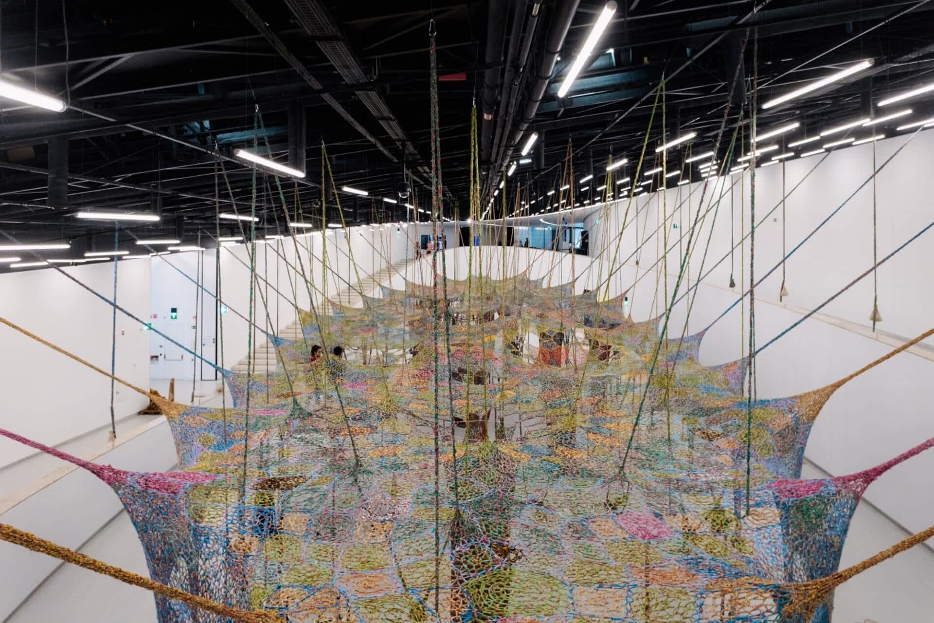 an overview of an expansive crocheted installation suspended in a large gallery space with rope-like pieces and weights draping it down to the floor