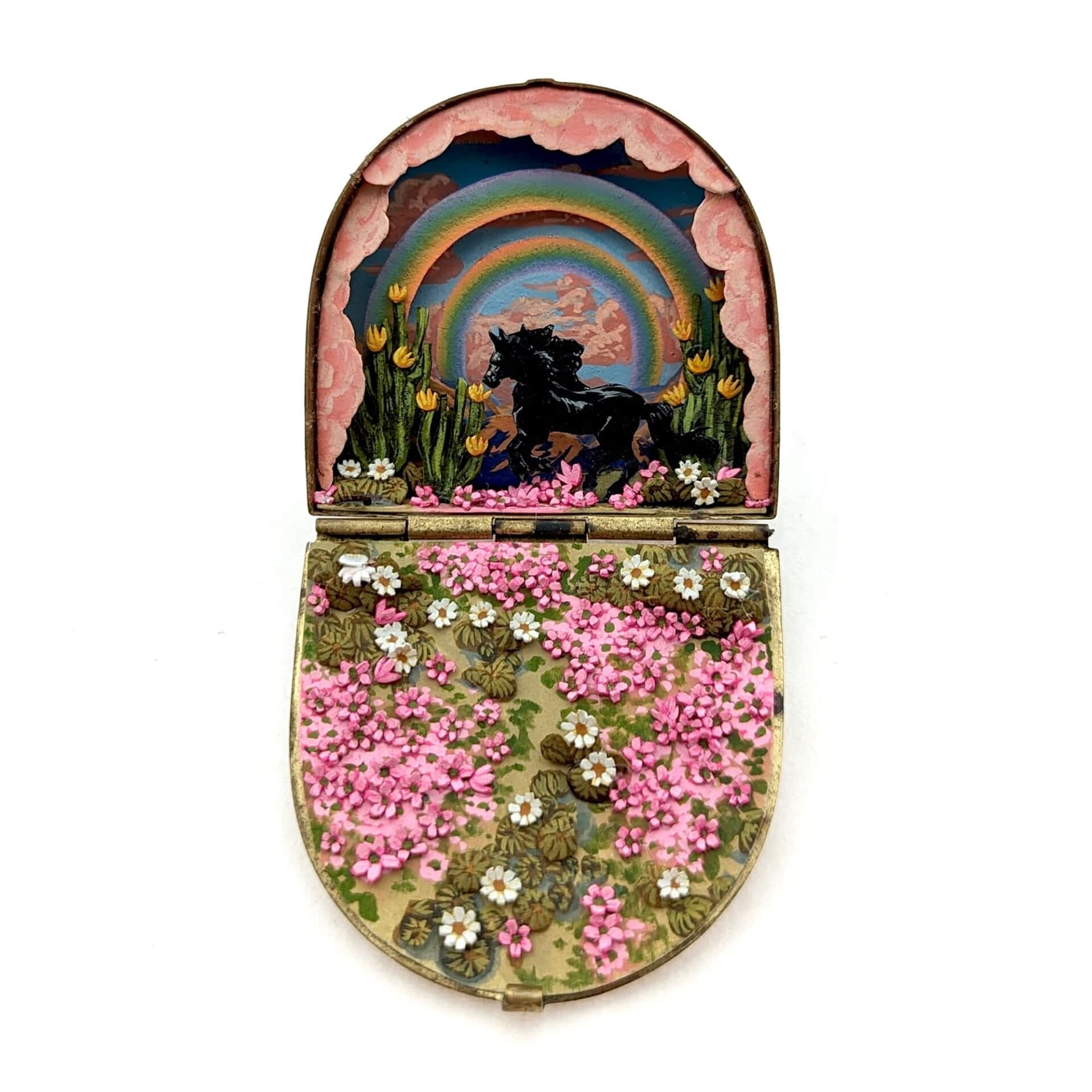 a tiny diorama made from watercolor-painted paper inside of a vintage cosmetics compact depicting a black stallion running underneath a double rainbow and pink clouds in a field of pink flowers