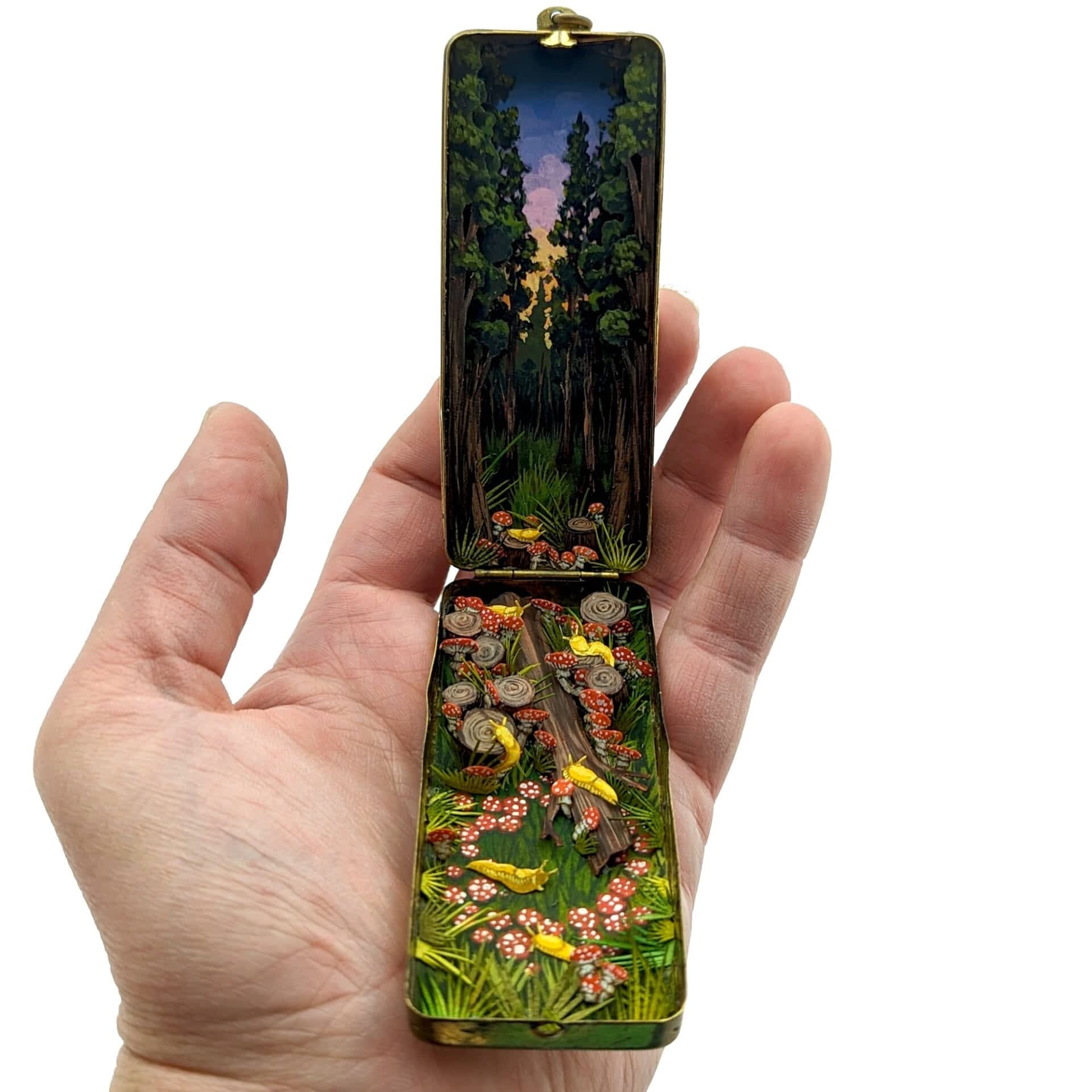 a tiny diorama held in a person's hand, made from watercolor-painted paper inside of a long vintage cosmetics compact depicting a woodland scene at dusk