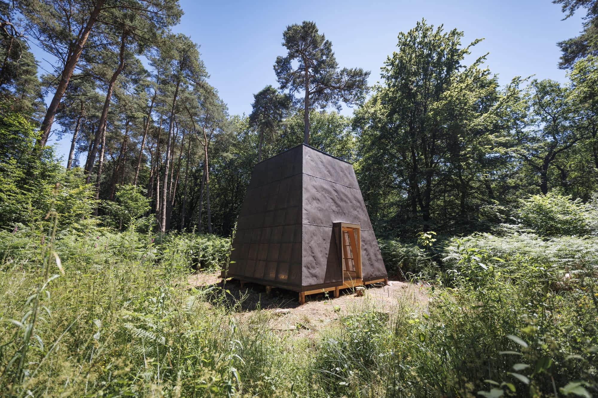 a geometric wooden building covered with black tarps stands in a forest