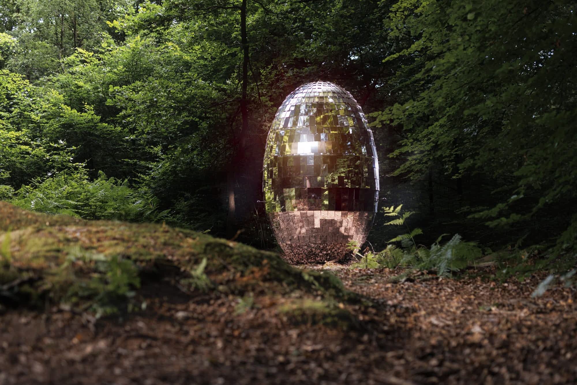 an ovoid shape covered in mirrored panels like a disco ball stands in the middle of a forest, shining in the sunlight