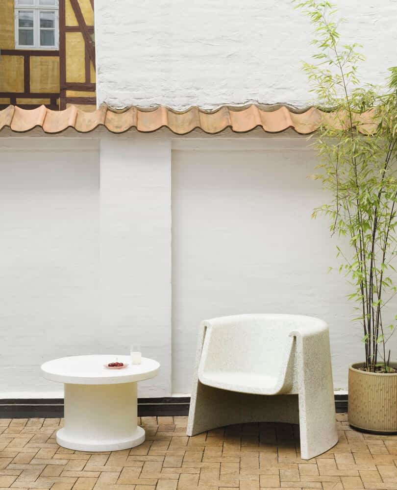 An outdoor seating area with a white armchair and round white table on a brick-paved surface, adjacent to a white wall with a clay-tile roof. A potted bamboo plant stands nearby.