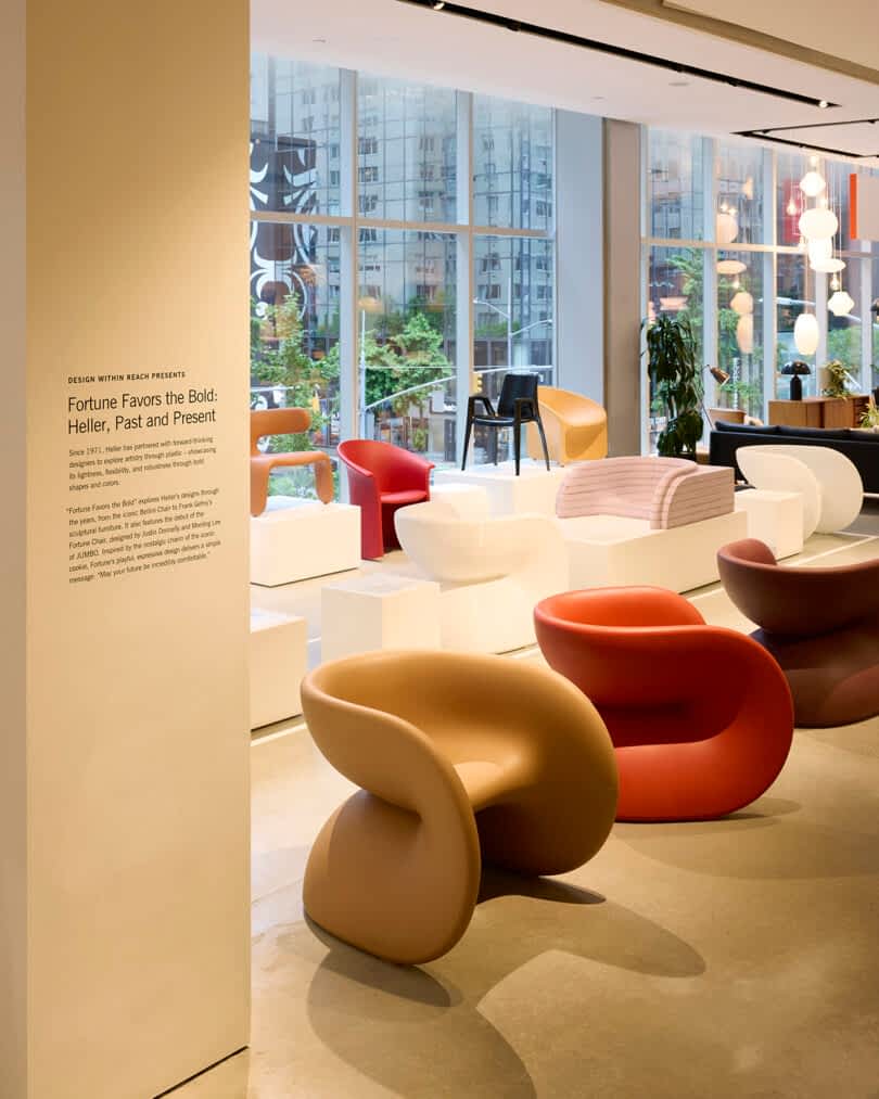 An exhibition room features modern designer chairs, including red, brown, and white pieces, displayed on white podiums in front of large windows with a cityscape view.