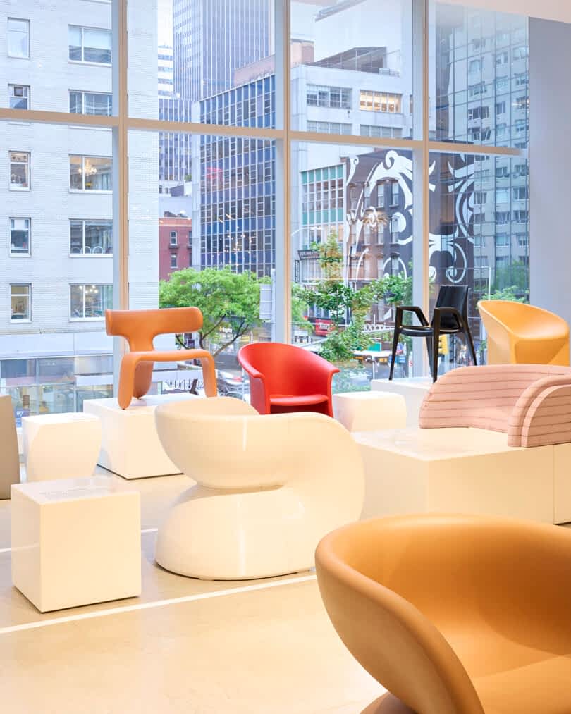 An exhibition room features modern designer chairs, including red, brown, and white pieces, displayed on white podiums in front of large windows with a cityscape view.