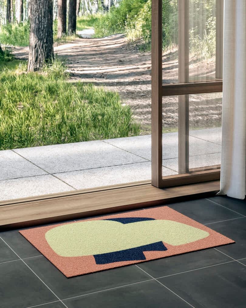 A colorful doormat placed by an open door, leading to a sunlit path.
