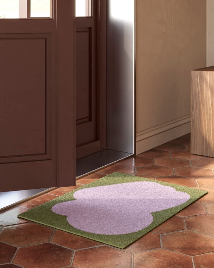 A lavender and green doormat placed on a tiled floor near an open wooden door.