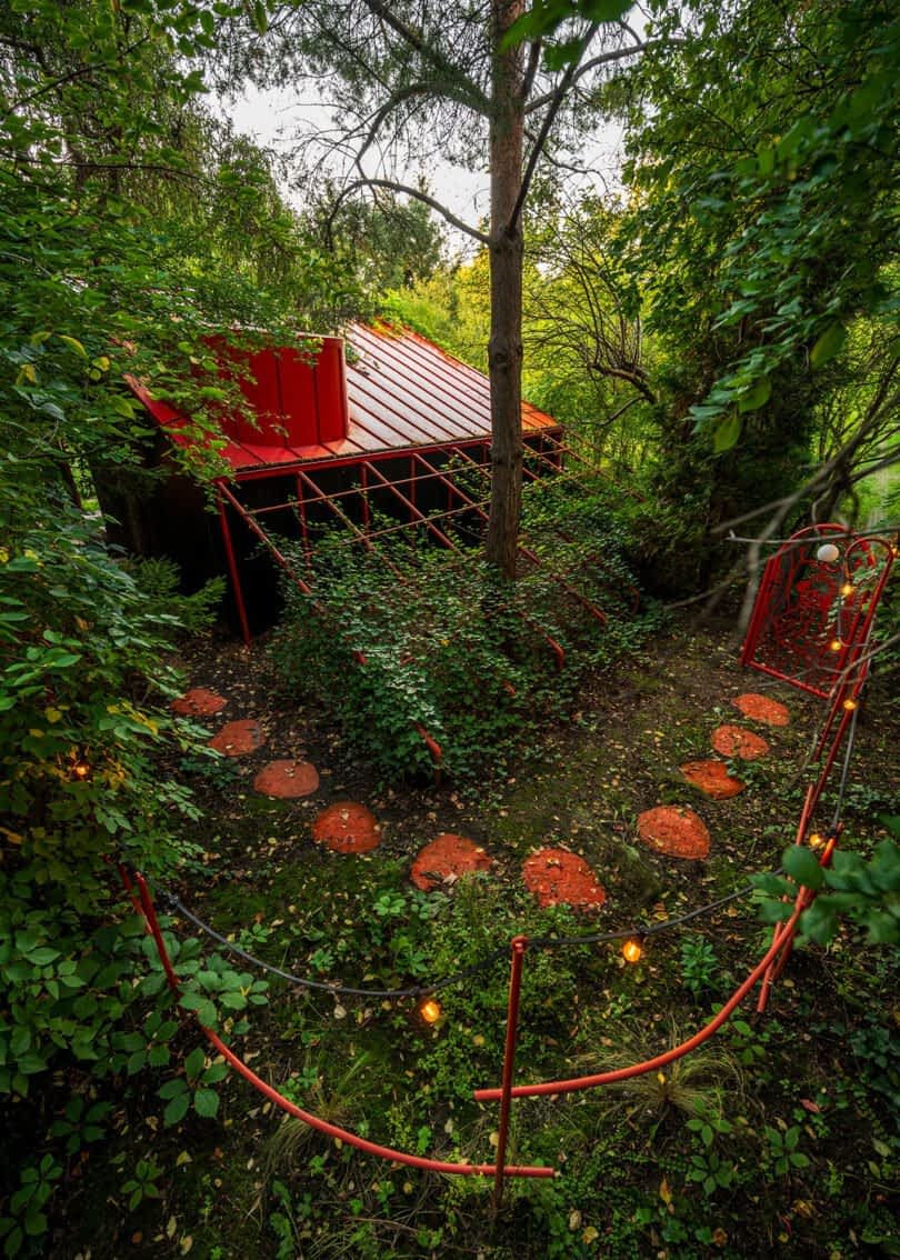 A small garden shed with a red roof is surrounded by lush greenery. It features a red metal gate and stepping stones leading up to it through the overgrown garden.