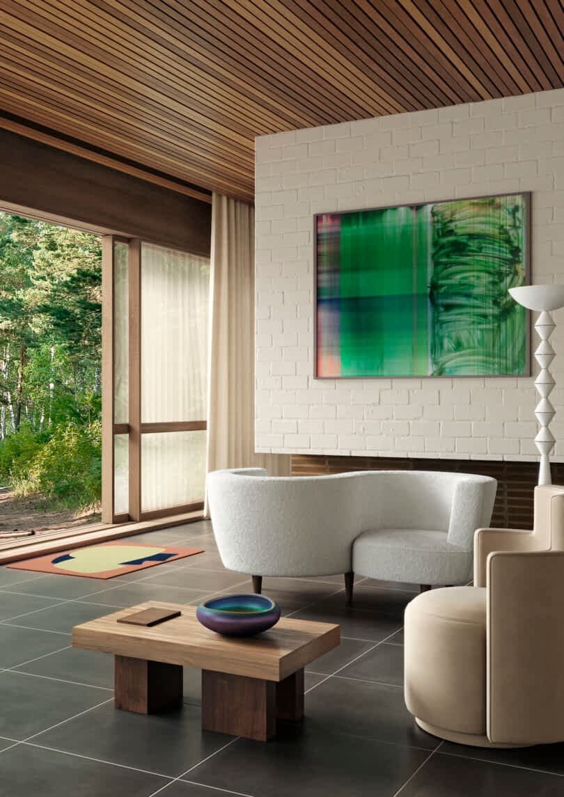 Modern living room with white sofa, beige armchair, and a wood coffee table, featuring large windows overlooking a forest, and an abstract painting on a white brick wall