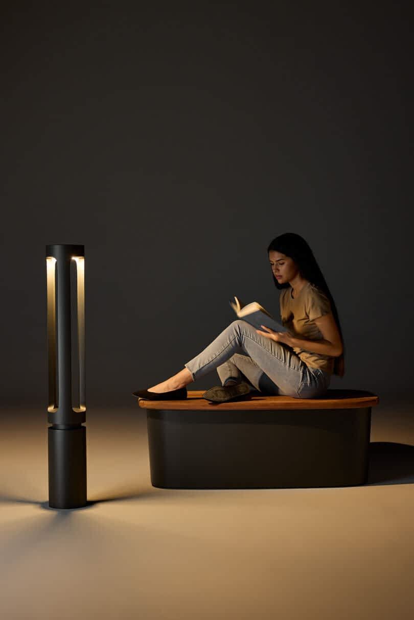 A woman with long hair sits reading a book in a dimly lit room. A tall, cylindrical lamp illuminates the scene.