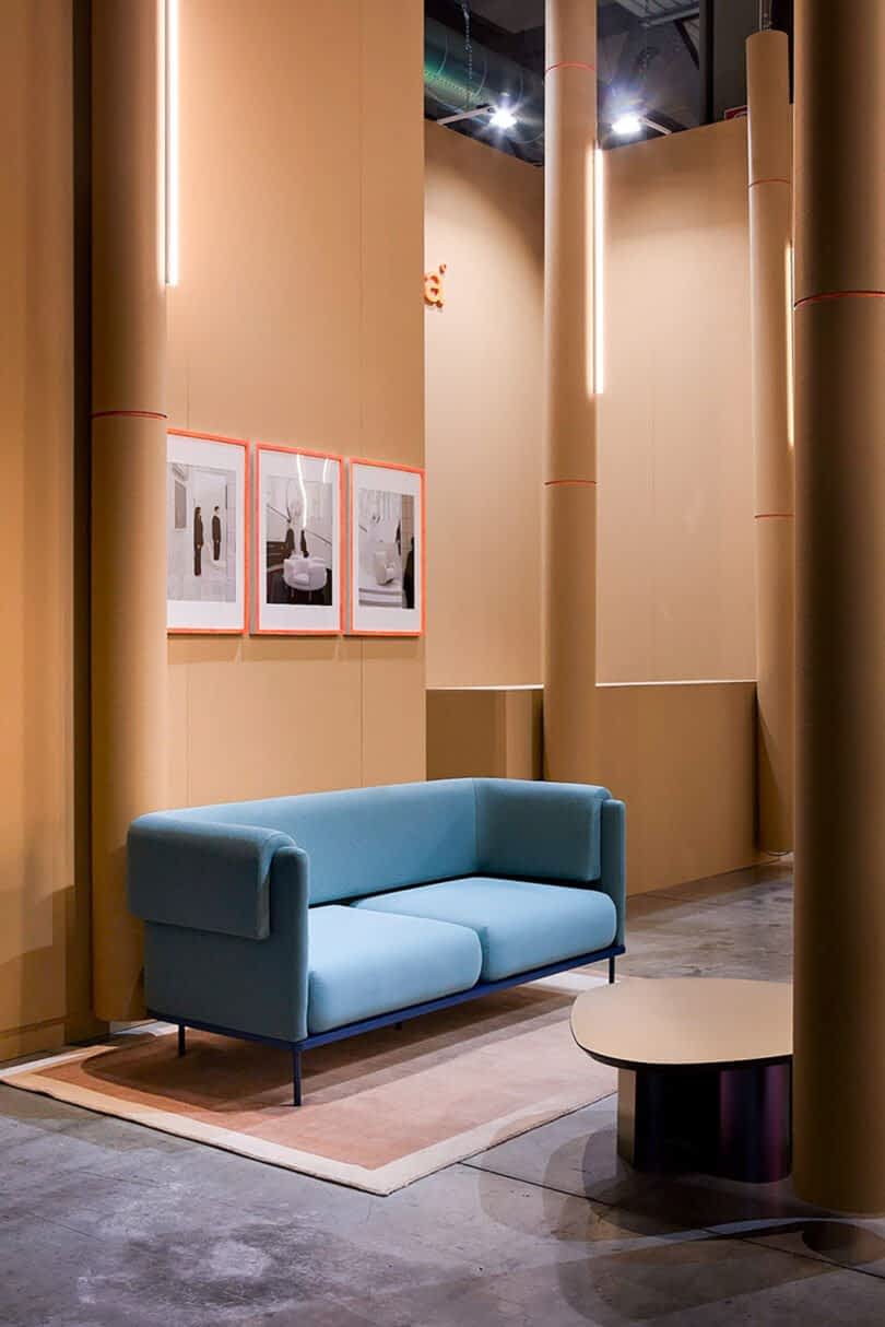 A modern showroom with a light blue sofa, round coffee table, beige walls, framed pictures, and vertical light fixtures.