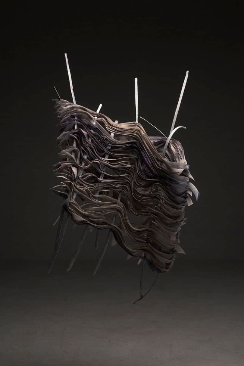 A sculptural artwork featuring layered, undulating dark material with protruding rods, displayed against a gray background.