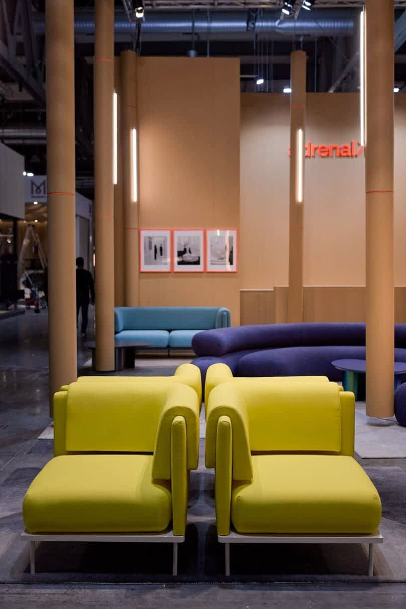 A modern showroom features yellow modular seating in the foreground, blue seating in the background, and framed artworks on a beige wall under industrial lighting.