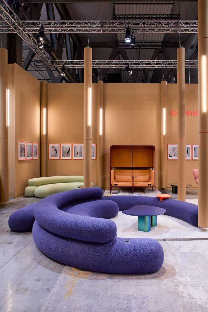 A modern showroom with curved purple seating, a central blue table, and various other colorful furniture pieces arranged on a concrete floor, surrounded by framed pictures on beige walls.