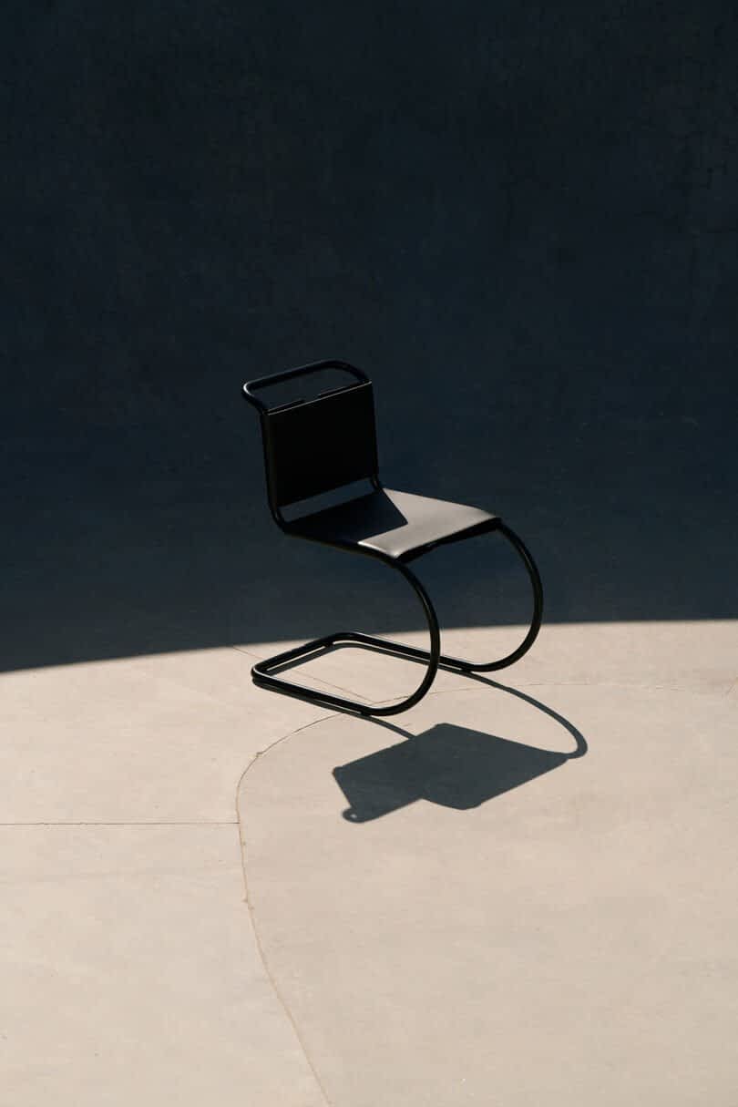 A modern black chair with a curved metal frame casting a shadow on a smooth concrete surface, partially illuminated by sunlight.