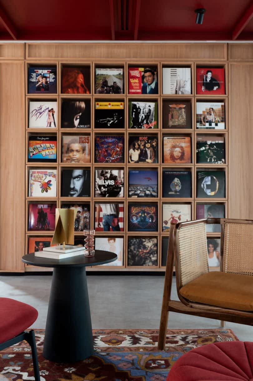 Wall display of vinyl records in a room with wooden shelves, a black round table, candle, and seating area with red and mustard-colored chairs