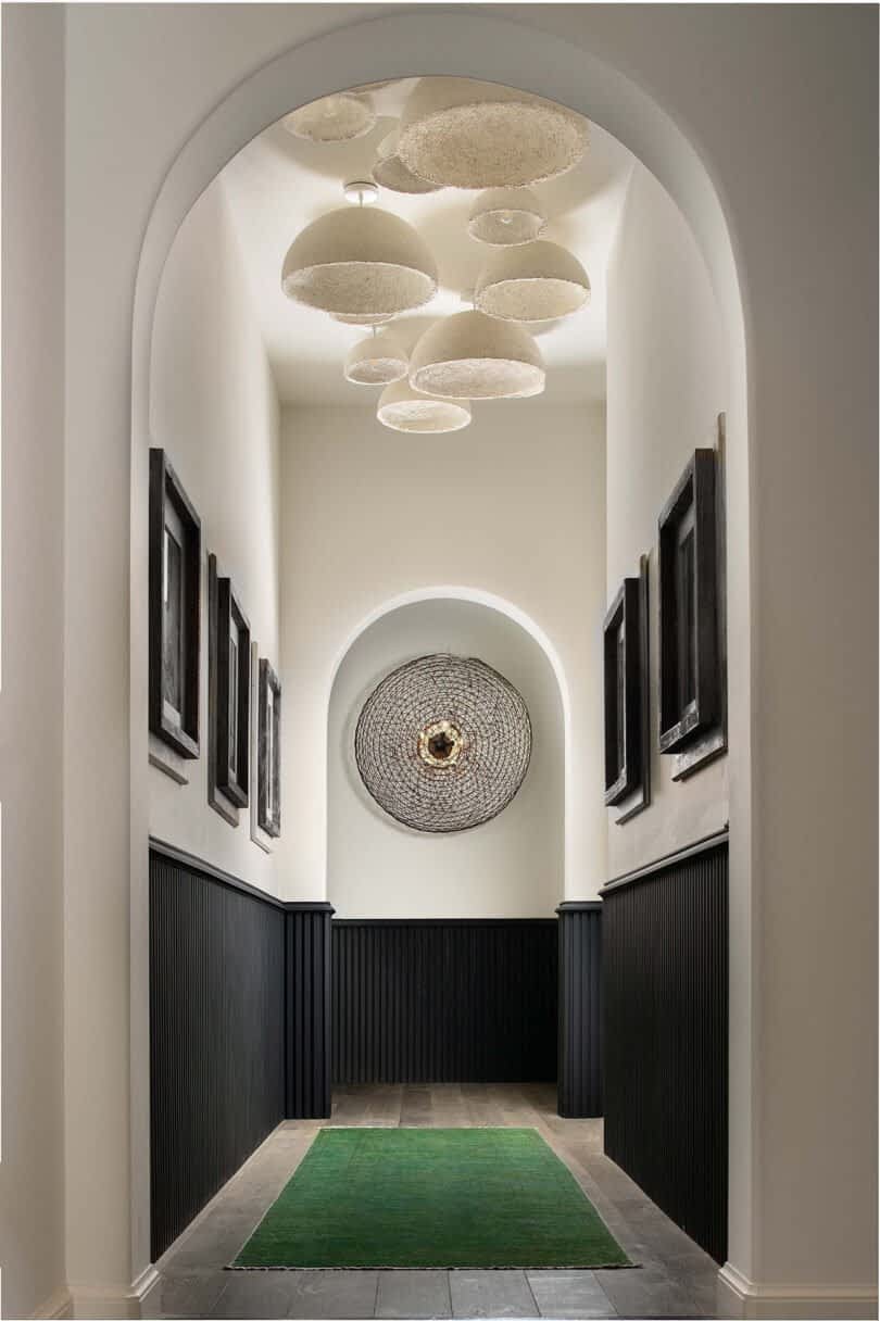 A narrow hallway with dark wainscoting, a green rug, framed pictures on the walls, and an array of MushLume Lighting decorative ceiling lights. At the end is a central arch with a large circular wall decoration.