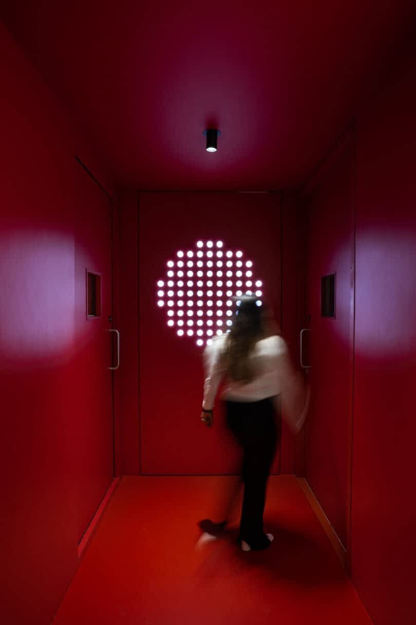 A person stands in a red-lit hallway with white round lights arranged in a circular pattern on the far wall. The person is blurred, indicating motion
