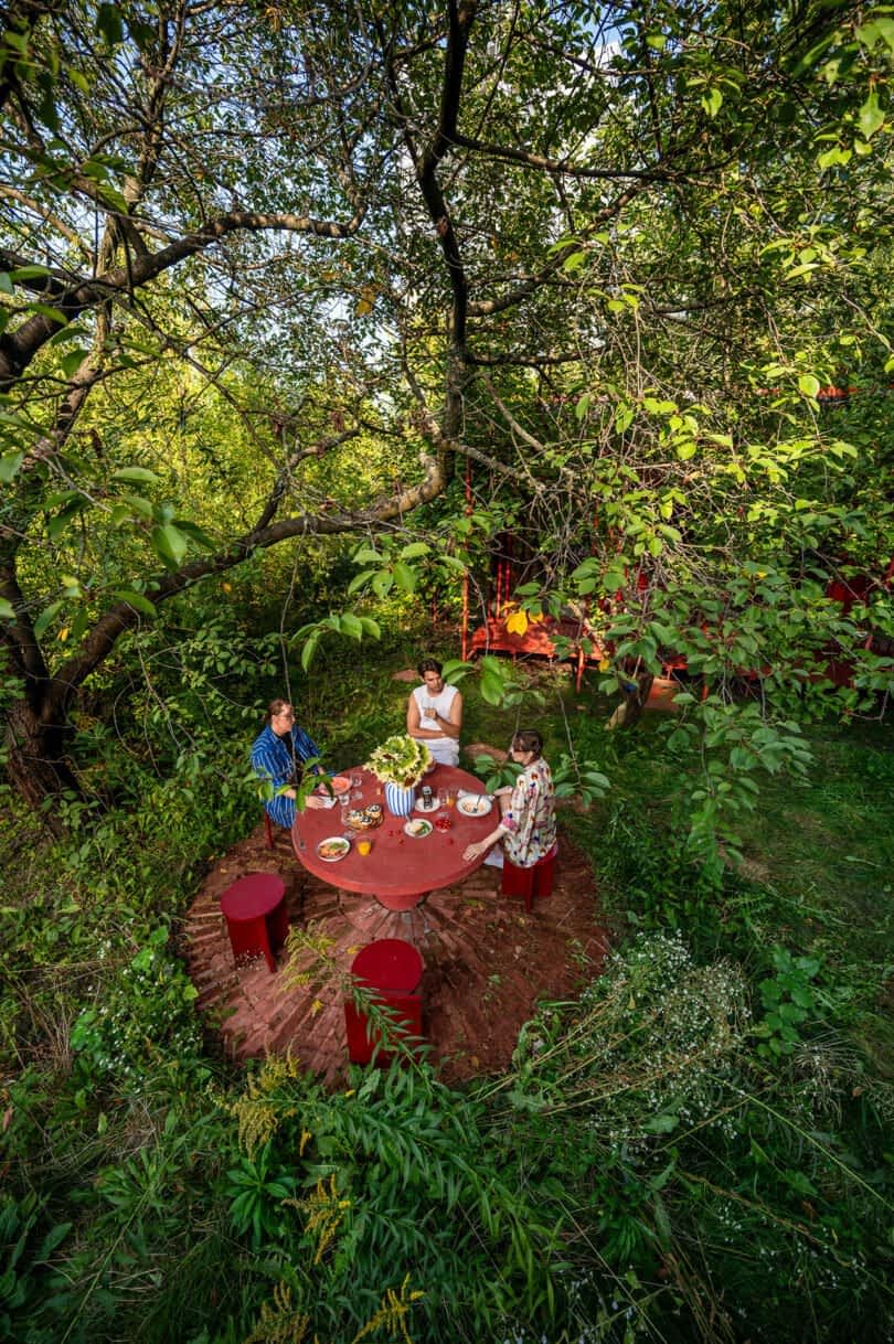 Four people sit at a round red table in a lush, green garden surrounded by trees and foliage, enjoying a meal together.