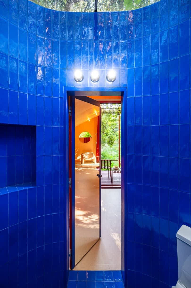 A doorway with blue tiles leads to a room with orange walls and a large window showing greenery outside. Three round lights are mounted above the door.