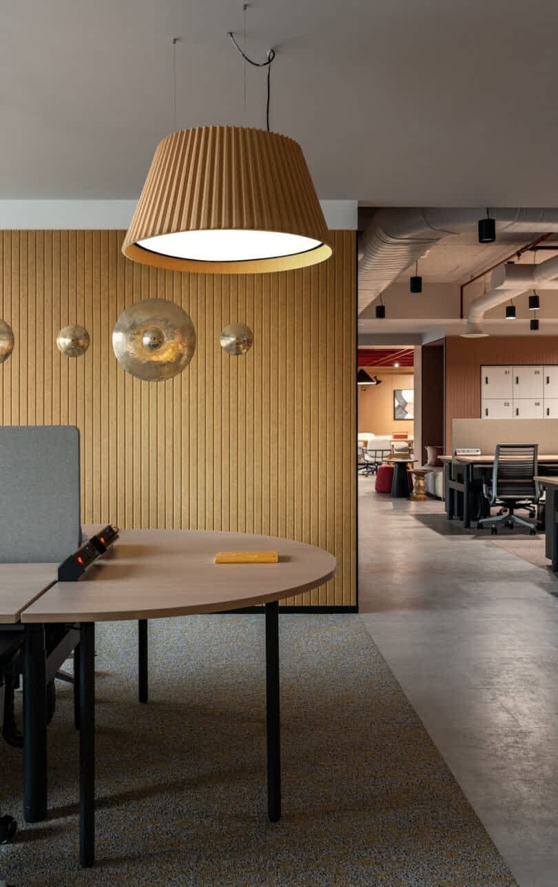 Modern office space with wooden paneling, round tables, and pendant lights. Several workstations and chairs are visible, with a mix of open and secluded seating areas