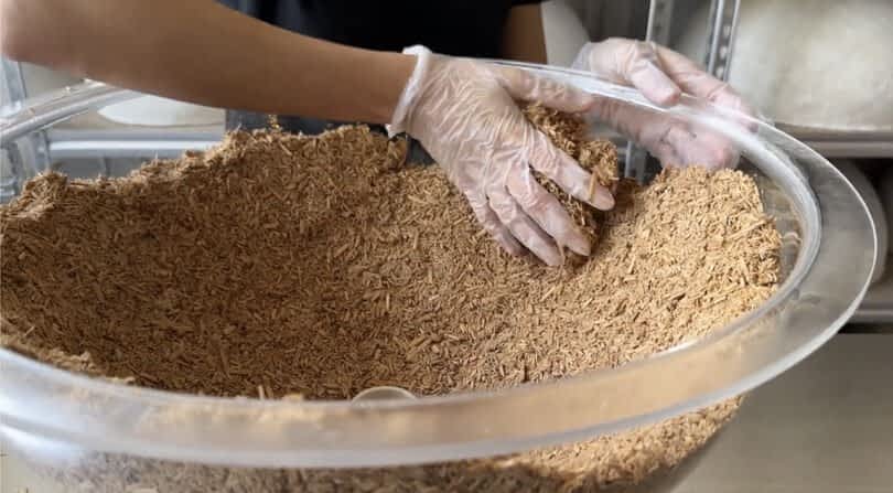 A person wearing gloves mixes a large transparent bowl filled with a shredded, brown material, perhaps for a MushLume Lighting project. Shelves are visible in the background.