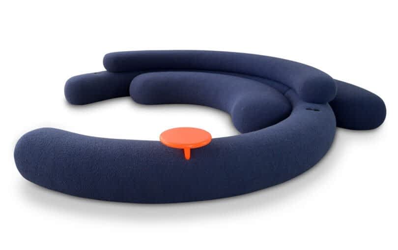 A dark blue, curved sofa with an orange side table attached to the middle section.