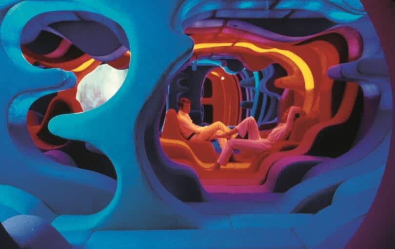 Two people in white outfits are sitting and talking in a futuristic, colorful, and abstractly designed room with vibrant lighting