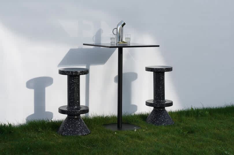 Two black speckled barstools and a square cafe table with two glasses and a pitcher are placed on a green lawn against a white wall.