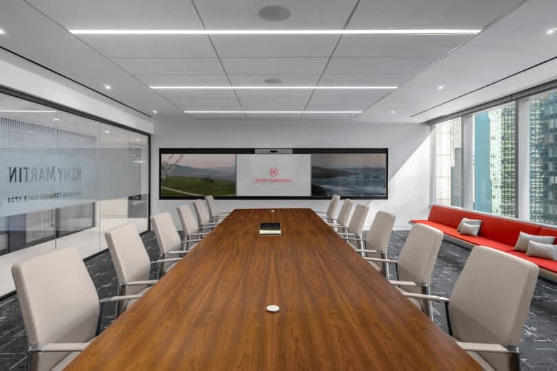 A modern conference room with a long wooden table, white chairs, a large screen displaying a logo, and floor-to-ceiling windows. A red couch with pillows is along one wall