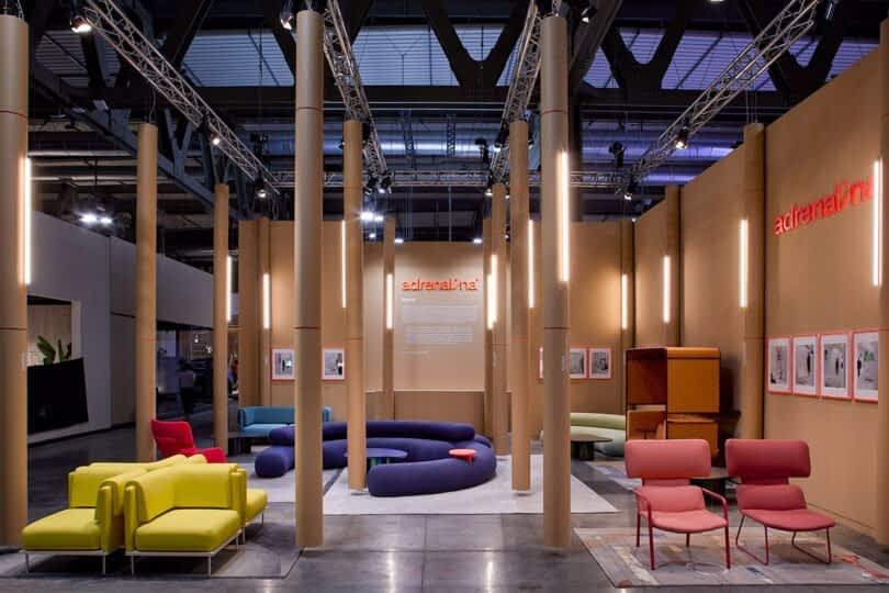 Modern furniture showroom with colorful chairs, couches, and a hanging orange sign with the word “adrenalina.” The booth is surrounded by tall light-colored columns under an industrial ceiling.