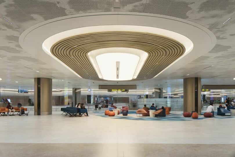 A spacious airport lounge with modern seating and a unique ceiling design, where travelers are seated and waiting.
