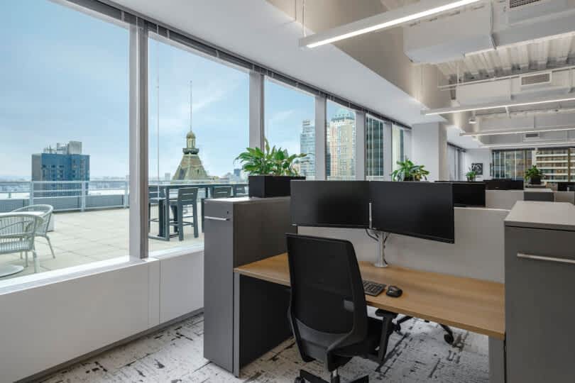 An empty office workspace with a desk, chair, and dual monitors, next to large windows overlooking a cityscape with buildings and a balcony with outdoor seating