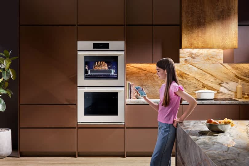Young girl in a pink shirt and jeans leaning against a kitchen island looking at her cell phone with the. SKS Transitional Double Wall Oven in the background. A roast is in the oven baking.