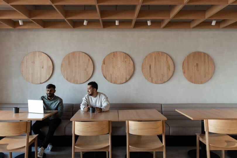 Two men sitting at a wooden table in a cafe, with one using a laptop and both holding mugs. The wall behind them features five circular wooden decorations