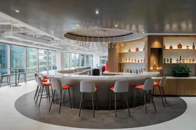 Modern bar area with a circular counter, red cushioned stools, and shelves of bottles against a backdrop of large windows overlooking city buildings. Ceiling features a contemporary light fixture