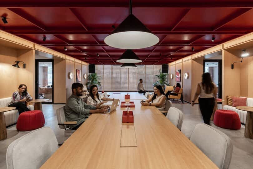 People are working in a modern office space with wooden furniture, red accents, and overhead lighting. Several individuals are seated at a long table with laptops, while others move about or sit in the background