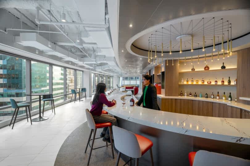 Two individuals are conversing at a modern, curved bar in a spacious, well-lit office lounge with large windows and an array of liquor bottles displayed on illuminated shelves