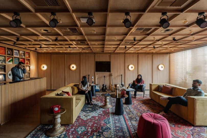 A modern, acoustically treated lounge with four people. The room features wooden paneling, a patterned rug, musical instruments, three yellow couches, and a mixing station with vinyl records on the wall