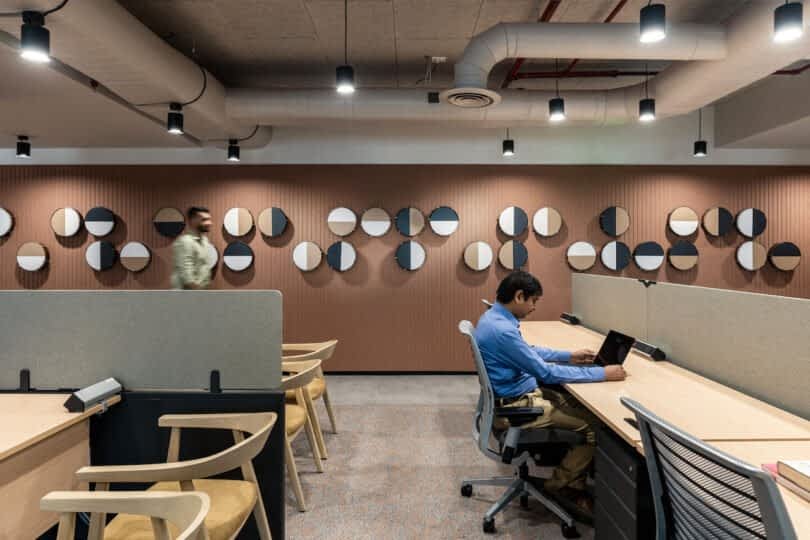 An office space with modern decor, including geometric wall art and ventilation ducts. One person is seated and working on a laptop, while another person walks by in the background