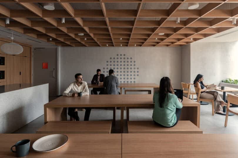 People are sitting and working on wooden tables in a modern, minimalist room with a grid ceiling, grey walls, and round wall art