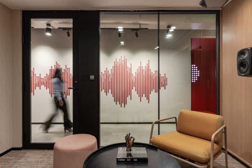 A modern office interior with a glass partition featuring a red soundwave design. A person is walking through a door on the left, and a seating area with a chair and ottoman is in the foreground