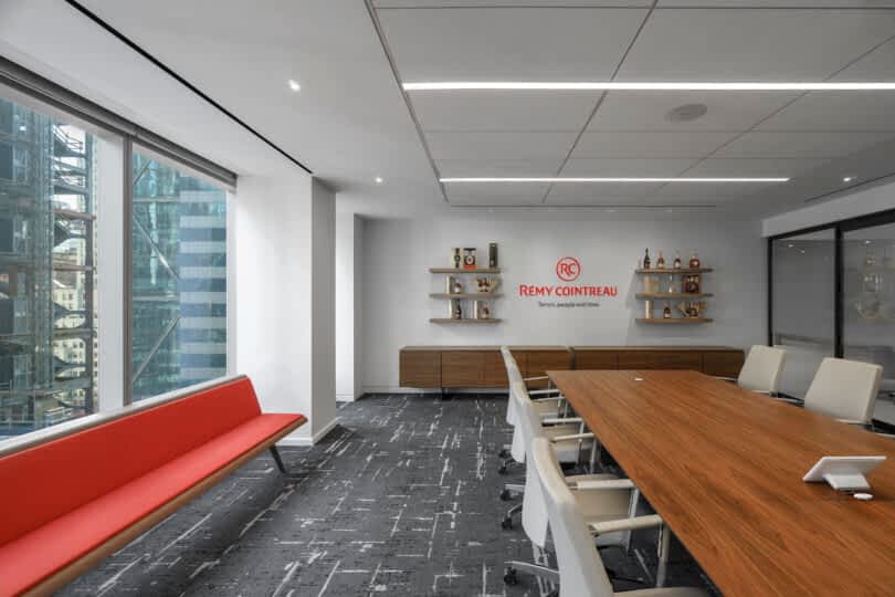 A modern office meeting room with a long wooden table, white chairs, a red bench, and a Remy Cointreau logo on the wall. Large windows provide a view of city buildings