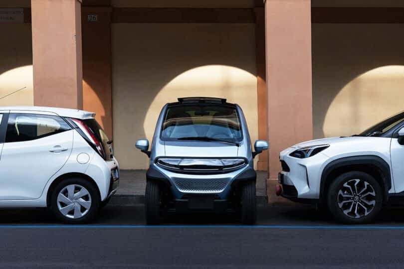 Three Eli ZERO micro EVs parked under an arcade, with sunlight casting circular shadows on the wall behind them.