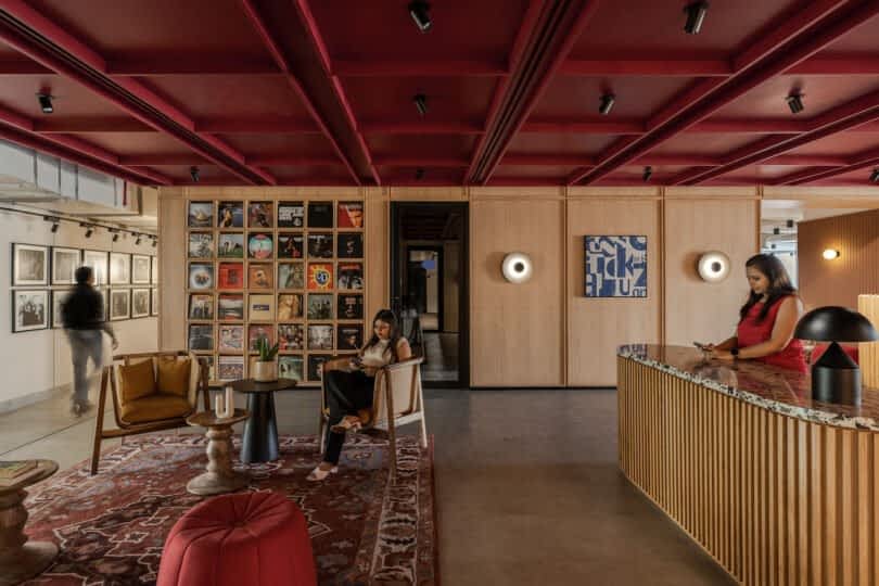 Modern office lobby with red ceiling, various artworks on walls, a seating area, and a reception desk where a woman stands using her phone. Another person sits in a chair, and a third walks by.