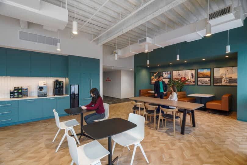 A modern break room with a kitchenette on the left, tables and chairs in the center, and a seating area with artwork on the wall in the background. Three people are present, one seated and two standing