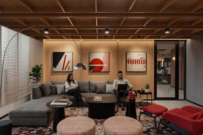 Two people sit in a modern office lounge with abstract artwork on the walls. One person uses a laptop while the other looks at their phone. The room features a sectional sofa and contemporary decor