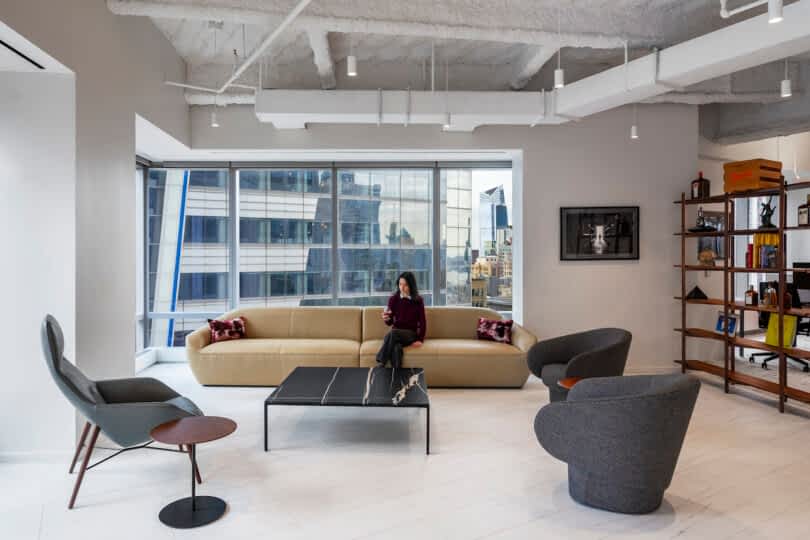 A person sits on a beige couch in a modern office with large windows, surrounded by contemporary furniture including gray chairs, a black coffee table, and a wooden bookshelf