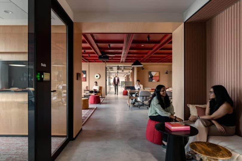 Modern office space with an open-plan design featuring wood and red accents. People are working and conversing on both sides of the corridor. Two women are sitting and chatting in the foreground
