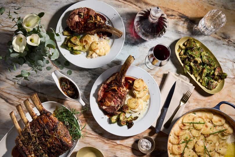A table spread of a roast with scalloped potatoes and Brussel sprouts, with wine and flowers decorating the table.