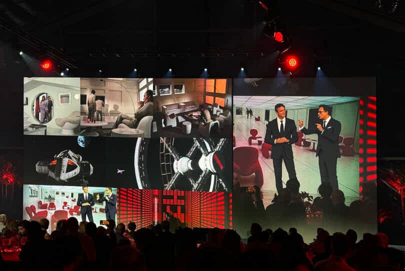 A large event screen displaying a variety of images including astronauts, spacecraft interiors, and two men conversing beside a Ferrari 12Cilindri sports car, viewed by an audience under red lighting.